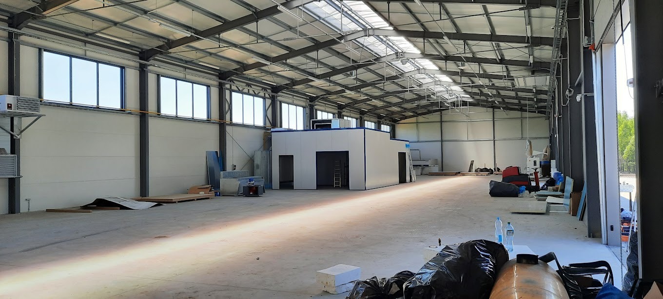 Wnętrze hali produkcyjnej z widoczną konstrukcją stalową dachu, oknami i instalacjami. Na podłodze materiały budowlane i sprzęt. Widoczne elementy instalacji elektrycznej.