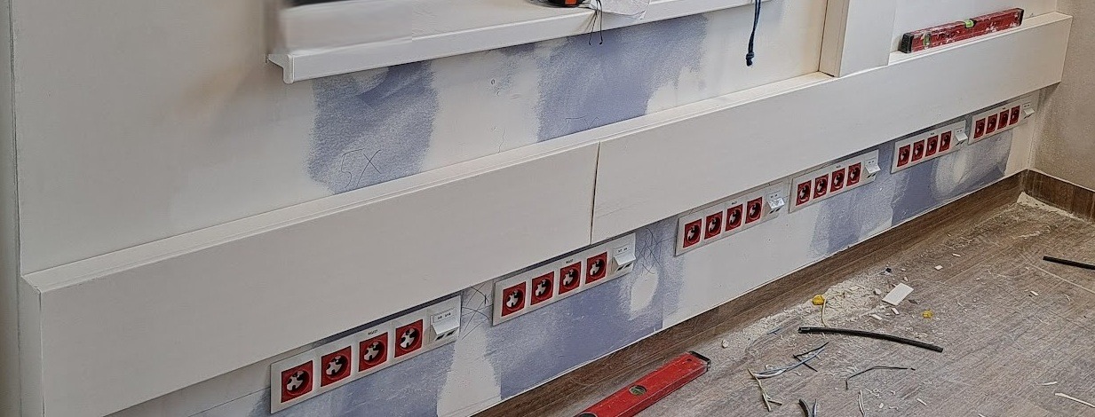 Rząd gniazd elektrycznych z czerwonymi wtykami, zamontowanych na ścianie wzdłuż białej listwy przypodłogowej. Widoczne ślady po pracach instalacyjnych i poziomica.
