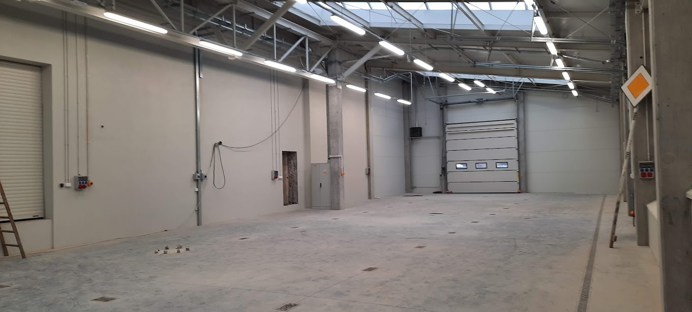 Wnętrze hali produkcyjnej z nową instalacją elektryczną: widoczne lampy, kable w listwach, skrzynki elektryczne na ścianach i brama segmentowa. Surowy beton i stalowa konstrukcja dachu.