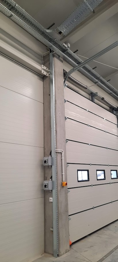 Biała brama segmentowa z oknami w hali przemysłowej. Widoczne szyny podsufitowe, betonowy słup z instalacją elektryczną i wyłącznikami.