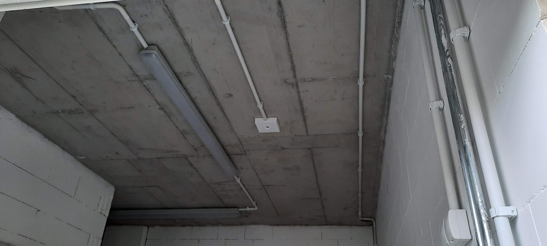 Szara, surowa betonowa powierzchnia sufitu z zamontowanymi białymi rurami instalacji elektrycznej i podłużnymi lampami. Widoczne uchwyty mocujące rury do sufitu.