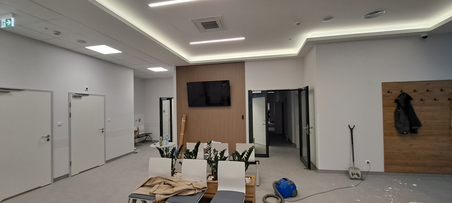 Nowoczesne wnętrze poczekalni z telewizorem na ścianie z drewnianymi panelami, oświetleniem LED i minimalistycznymi krzesłami. Widoczne ślady po remoncie.