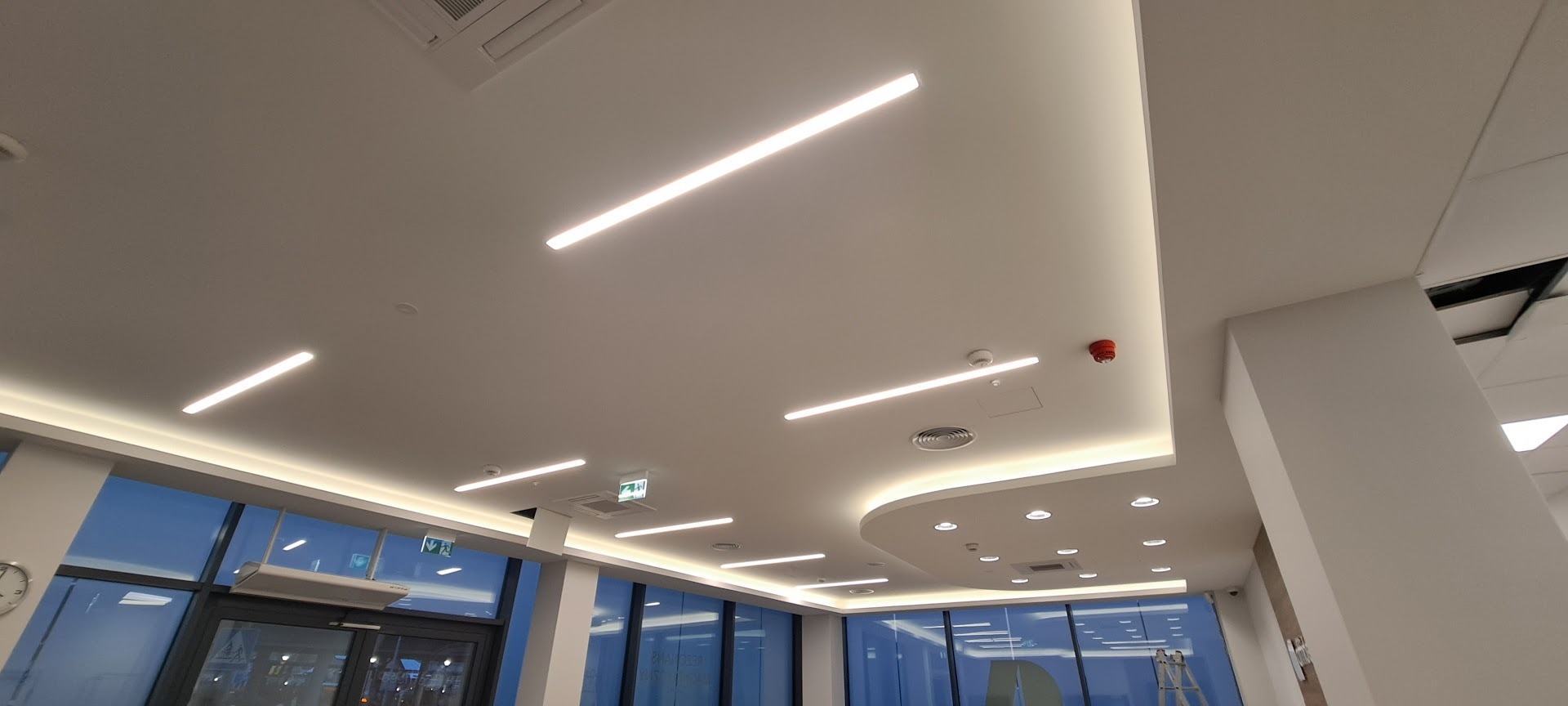 Biały sufit z liniowym oświetleniem LED w nowoczesnym biurze. Widoczne lampy, czujnik dymu i fragment okna z widokiem na zewnątrz. Minimalistyczny design.