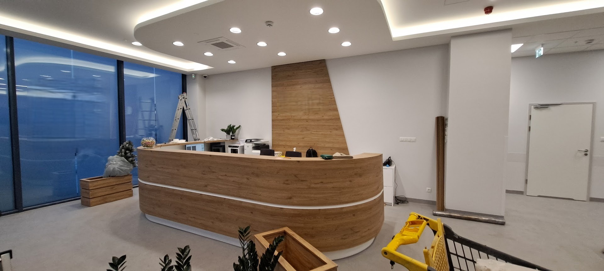 Nowoczesne wnętrze biurowe z drewnianą ladą recepcyjną, oświetleniem sufitowym i dużymi oknami. Widoczna drabina i elementy wykończeniowe sugerują prace w toku.