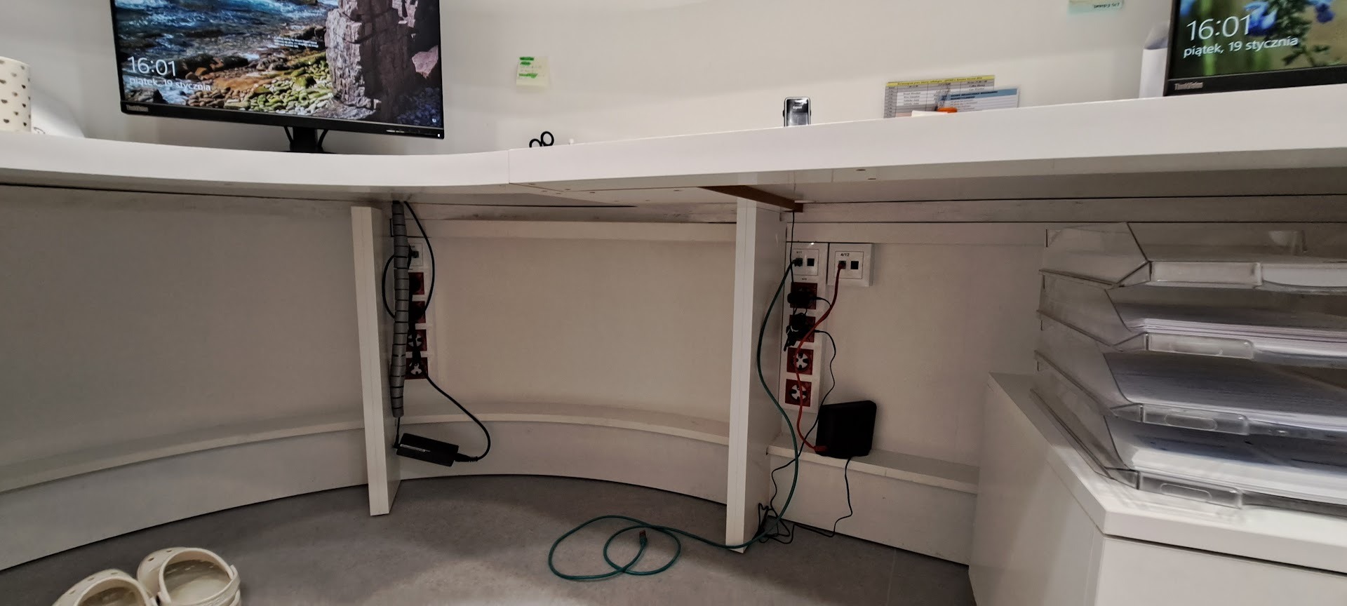 Ukryte gniazdka i kable pod biurkiem z monitorem. Listwa zasilająca zamontowana pionowo. Estetyczne rozwiązanie dla uporządkowania przestrzeni roboczej.