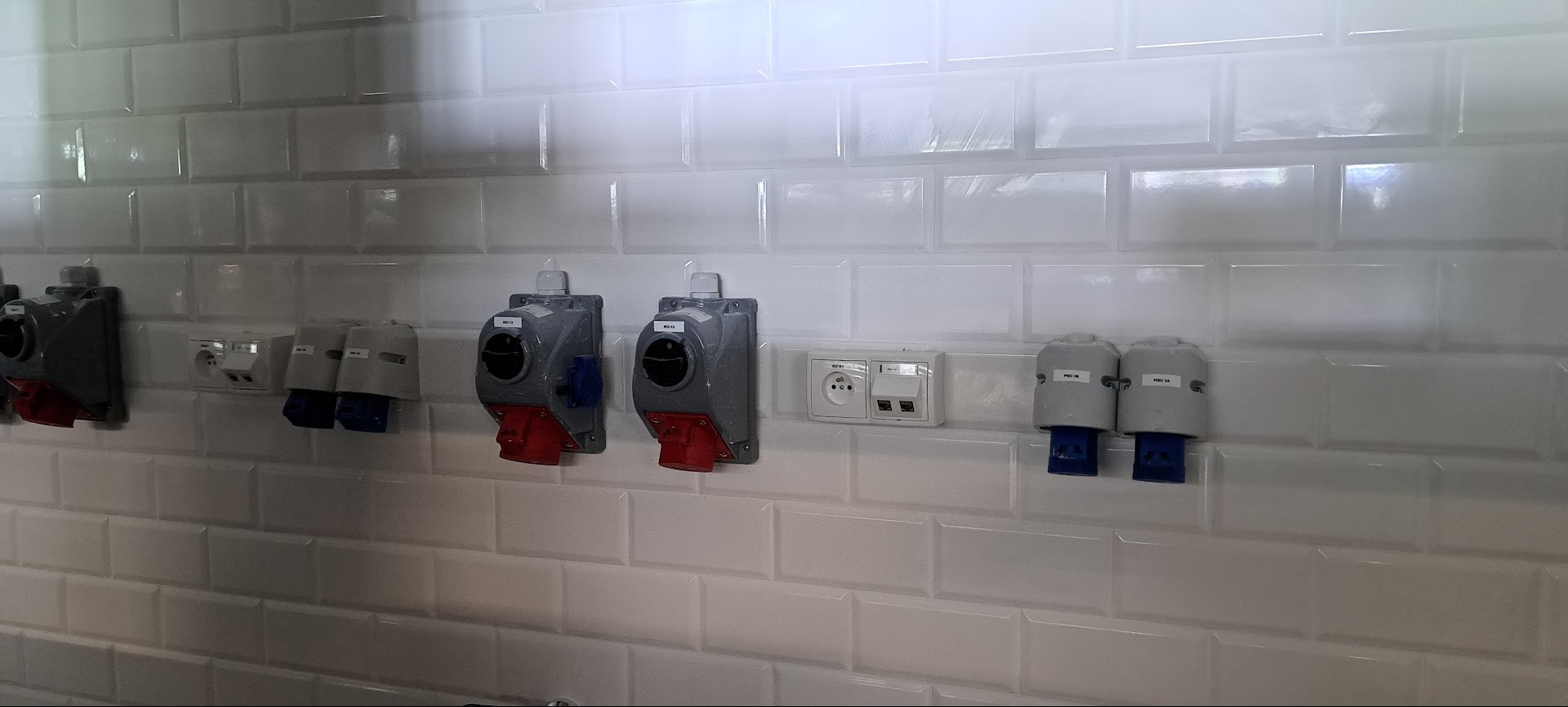 Rząd gniazd przemysłowych i standardowych na białej, kafelkowej ścianie. Widoczne różne typy gniazd, w tym gniazda siłowe z czerwonymi i niebieskimi elementami.