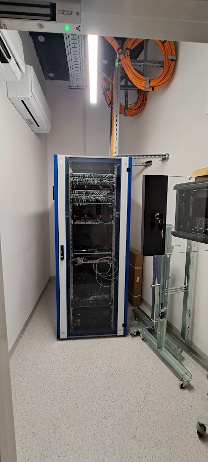 Szafa rack z okablowaniem w serwerowni, widoczne kable pomarańczowe na ścianie, klimatyzacja i oświetlenie. Sprzęt sieciowy w szafie.