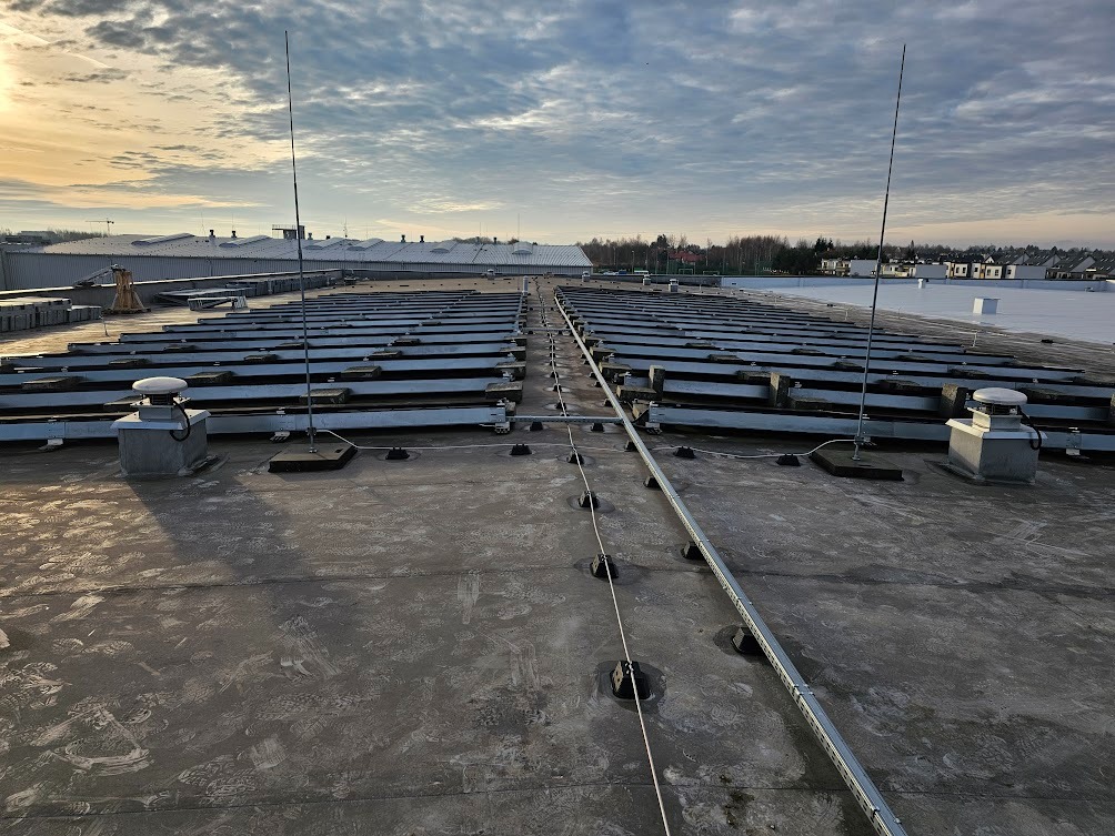 Instalacja odgromowa na dachu budynku przemysłowego z widocznymi masztami i przewodami uziemiającymi, w tle zabudowa miejska i pochmurne niebo.