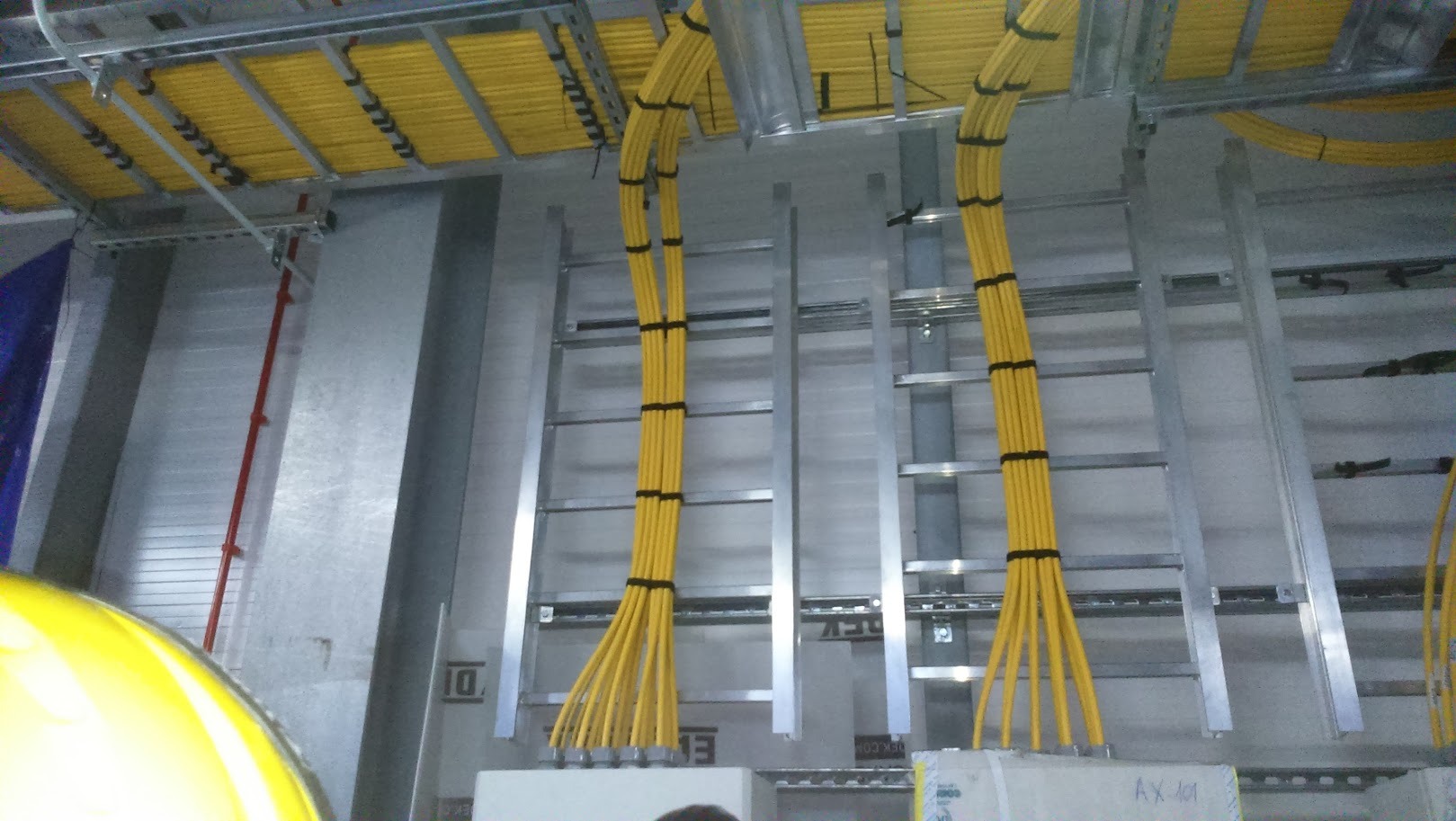 Żółte kable elektryczne ułożone w korytach kablowych, przymocowane czarnymi opaskami, widoczne elementy konstrukcji metalowej i fragment kasku ochronnego.