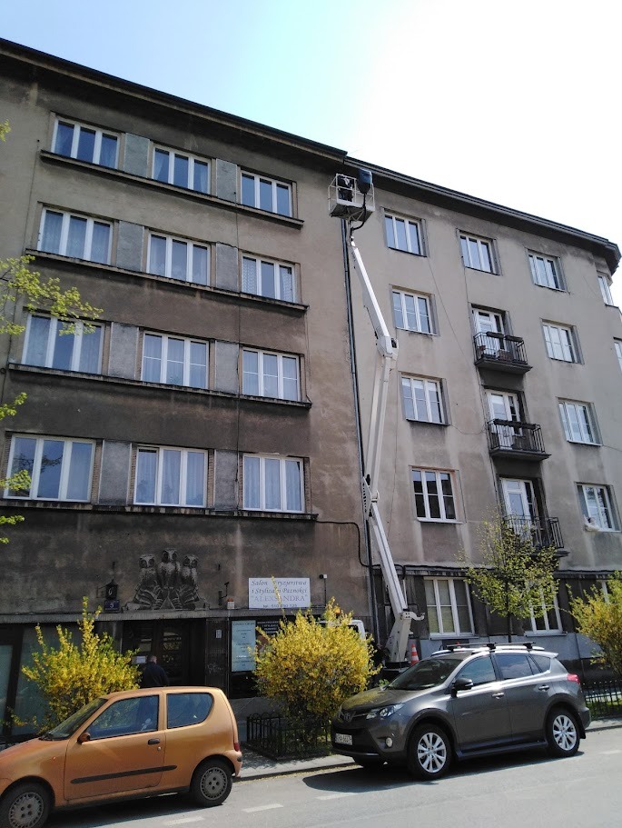 Prace na wysokościach przy budynku mieszkalnym z użyciem podnośnika koszowego. Widoczne okna, elewacja i zaparkowane samochody. Dzień słoneczny.