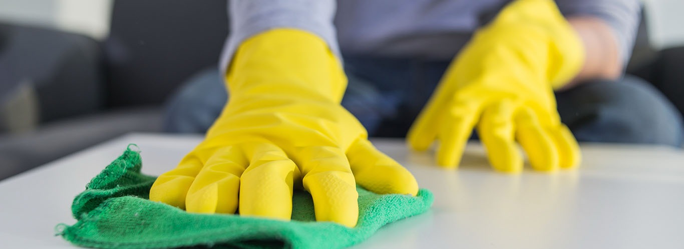 Dłoń w żółtej rękawicy ochronnej czyści białą powierzchnię zieloną ściereczką z mikrofibry; w tle zamazane siedzenie krzesła.