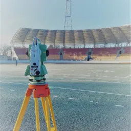 Teodolit Leica TS06 plus na trójnogu na tle stadionu sportowego z pustymi trybunami i masztem oświetleniowym w słoneczny dzień.