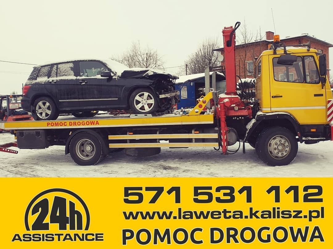 Żółta laweta z uszkodzonym czarnym SUV-em na platformie, częściowo pokrytym śniegiem, na tle zaśnieżonego terenu i budynku z czerwonej cegły.