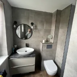 Nowoczesna łazienka w szarościach z drewnopodobną podłogą, owalną umywalką na szafce i okrągłym lustrem z odbiciem fotografa.