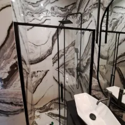 Nowoczesna łazienka z kabiną prysznicową typu walk-in z czarnymi profilami, umywalką nablatową o nietypowym kształcie i marmurowymi płytkami na ścianach.