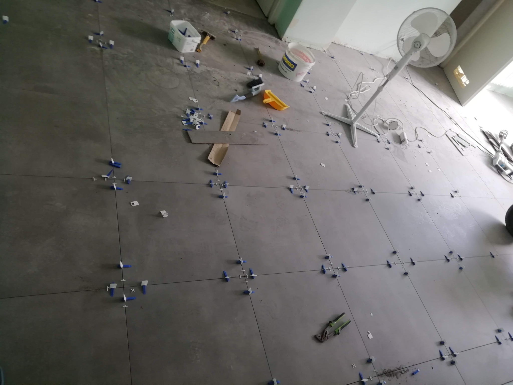 Szare płytki podłogowe w trakcie układania, widoczne krzyżyki dystansowe, narzędzia glazurnicze, wiadro z klejem oraz stojący wentylator w tle