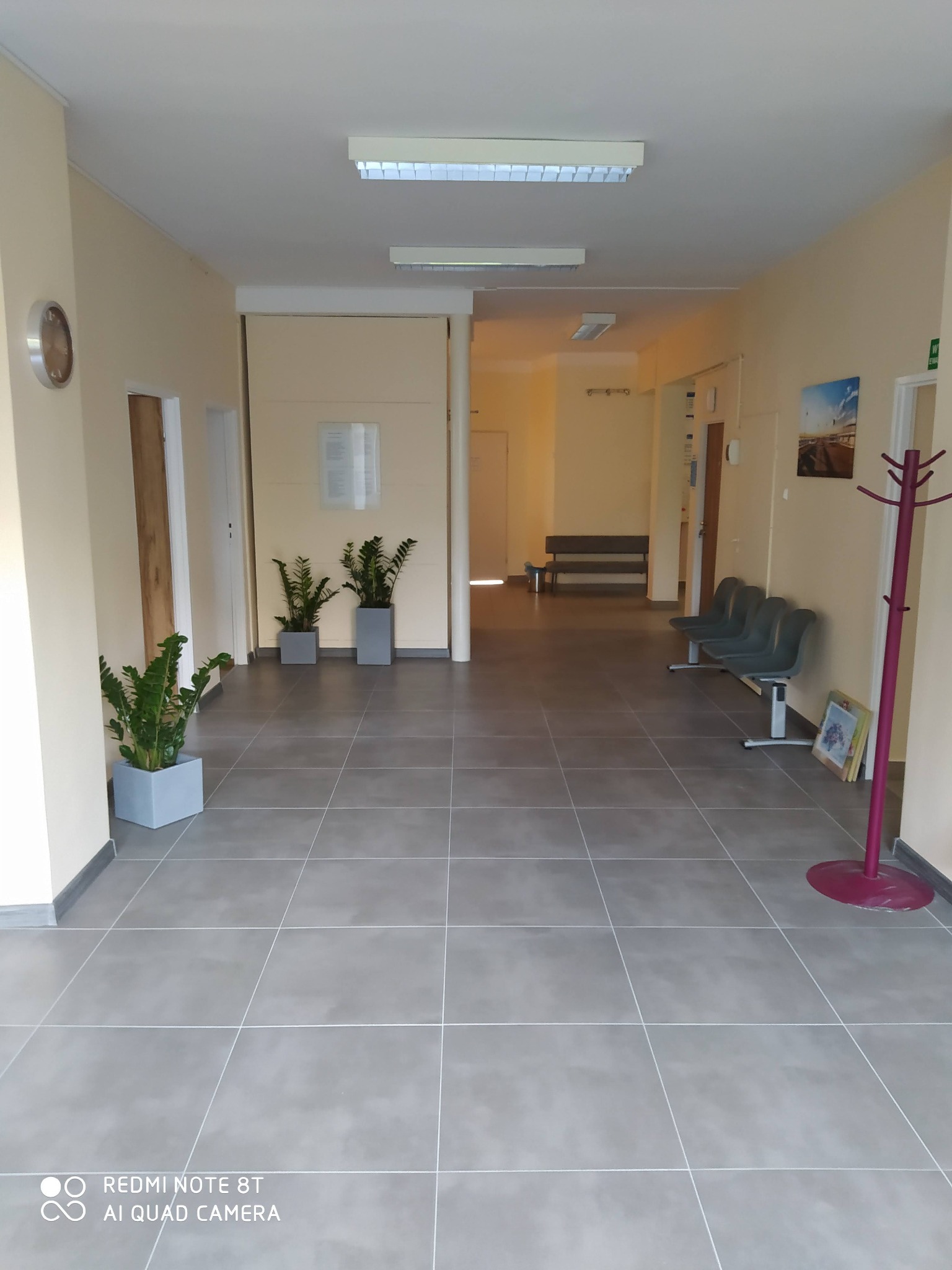 Długi korytarz wyłożony szarymi, kwadratowymi płytkami podłogowymi, z roślinami w donicach, krzesłami dla oczekujących i wieszakiem na ubrania, oświetlony lampami sufitowymi.