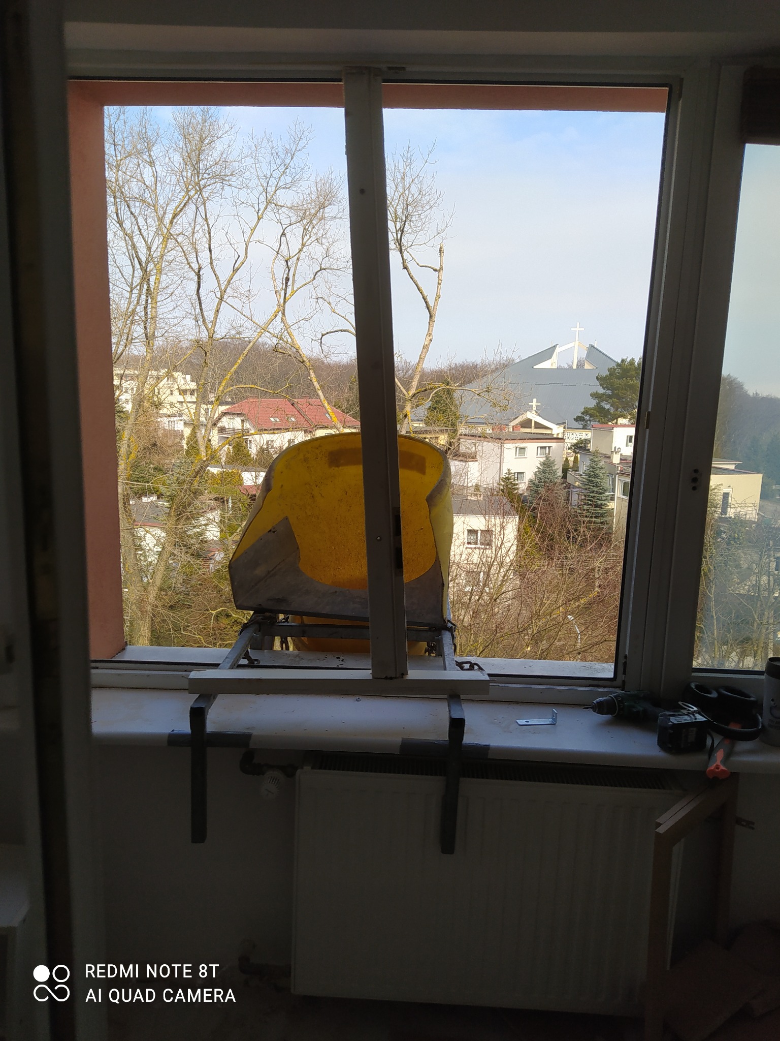 Żółty pojemnik na gruz umieszczony na parapecie okna podczas prac remontowych, widok na budynki mieszkalne i kościół w tle.