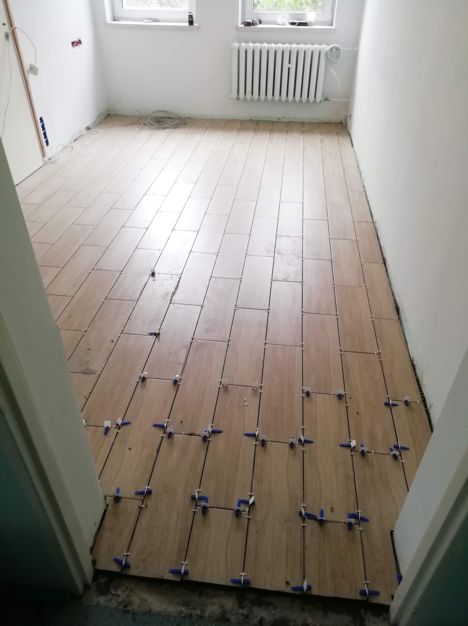 Jasne płytki podłogowe ułożone w pomieszczeniu, z widocznymi niebieskimi krzyżykami dystansowymi, białe ściany i grzejnik w tle, widok zza progu.