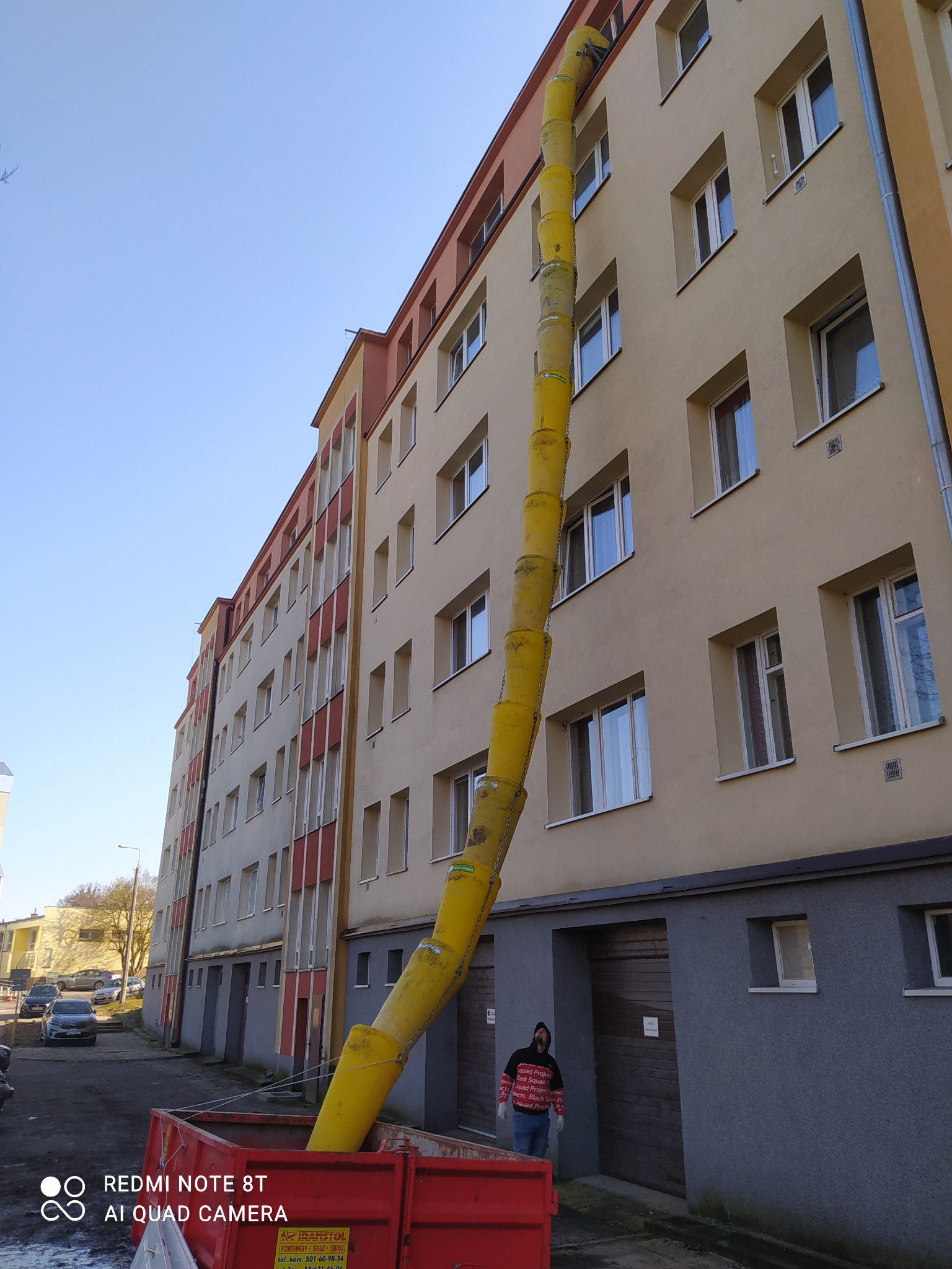 Żółty zsyp budowlany przymocowany do budynku wielorodzinnego, prowadzący do czerwonego kontenera na gruz, z pracownikiem w pobliżu.