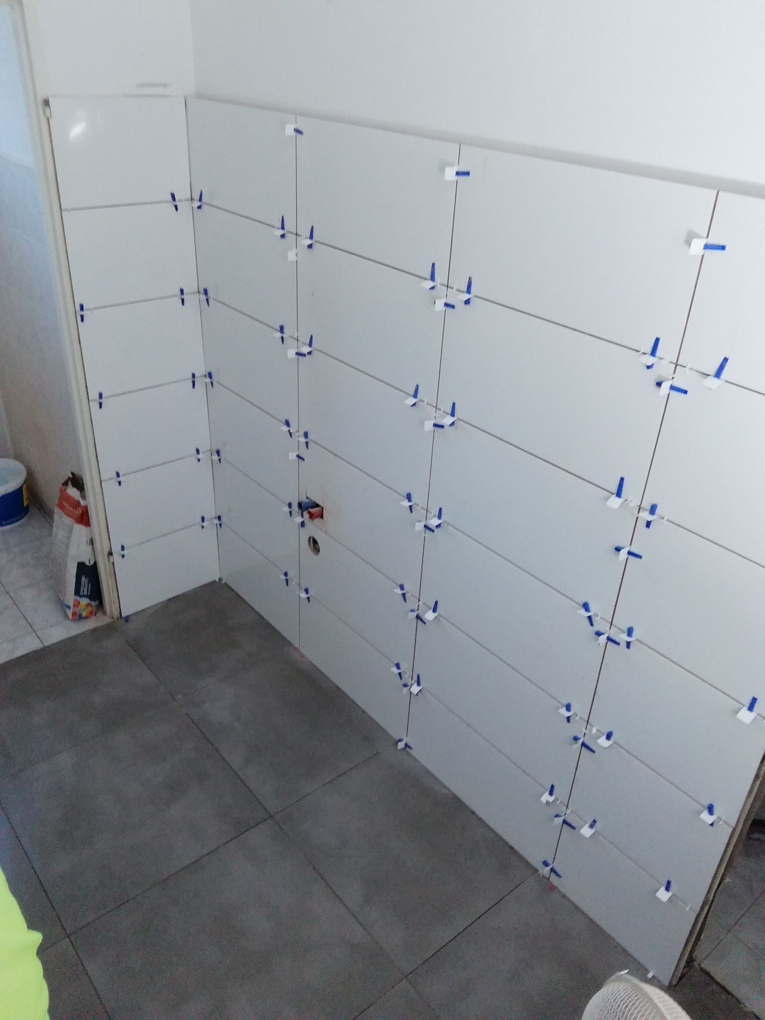 Świeżo ułożone białe płytki ścienne z niebieskimi krzyżykami dystansowymi, szare płytki podłogowe, widoczna instalacja wodna w ścianie, torba z zaprawą w rogu pomieszczenia.