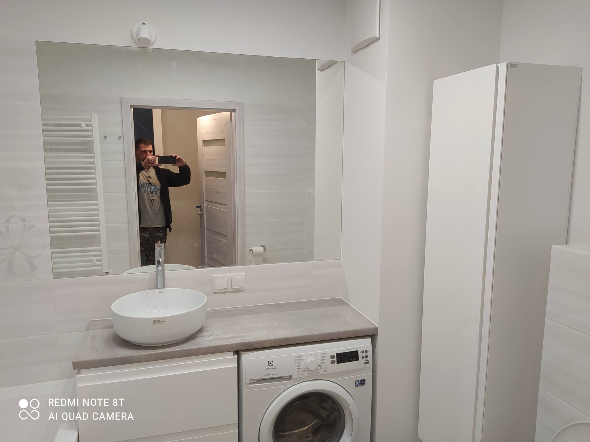 Nowoczesna łazienka w bieli z umywalką nablatową, pralką Electrolux i dużym lustrem, w którym odbija się osoba robiąca zdjęcie.