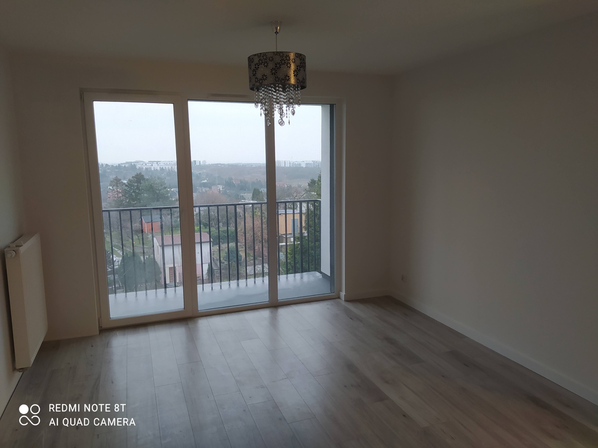 Pusty pokój z panelami podłogowymi w odcieniach szarości, dużym oknem balkonowym z widokiem na miasto i ozdobnym żyrandolem z kryształkami.