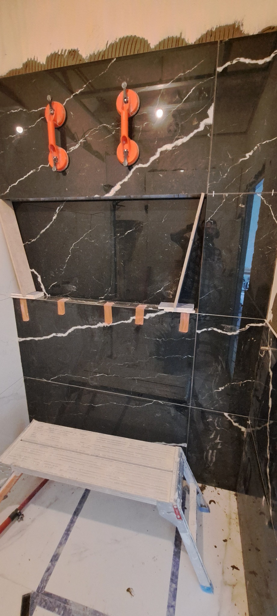 Montaż czarnych płytek imitujących marmur z białymi żyłami na ścianie, widoczne przyssawki do płytek i poziomica, drabina malarska w dolnej części kadru.