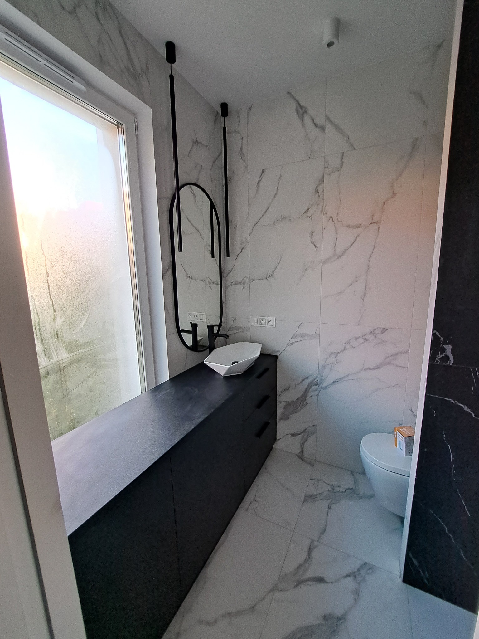 Nowoczesna łazienka z marmurowymi płytkami, czarną szafką pod umywalką, owalnym lustrem i wiszącą toaletą.