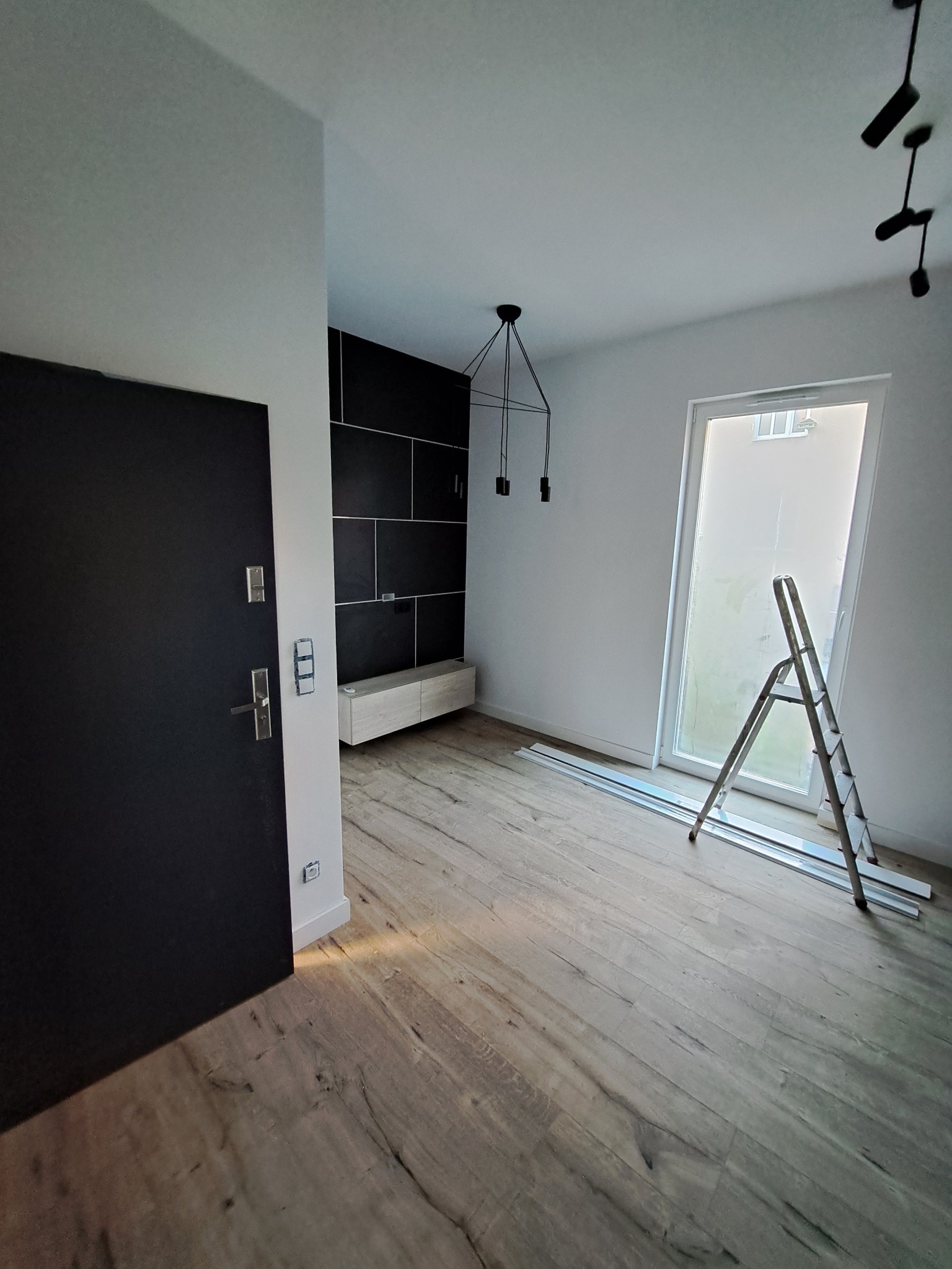 Wnętrze pokoju w trakcie wykańczania: drewnopodobna podłoga, czarne płytki na ścianie, drabina oparta o okno, listwy przypodłogowe na podłodze.