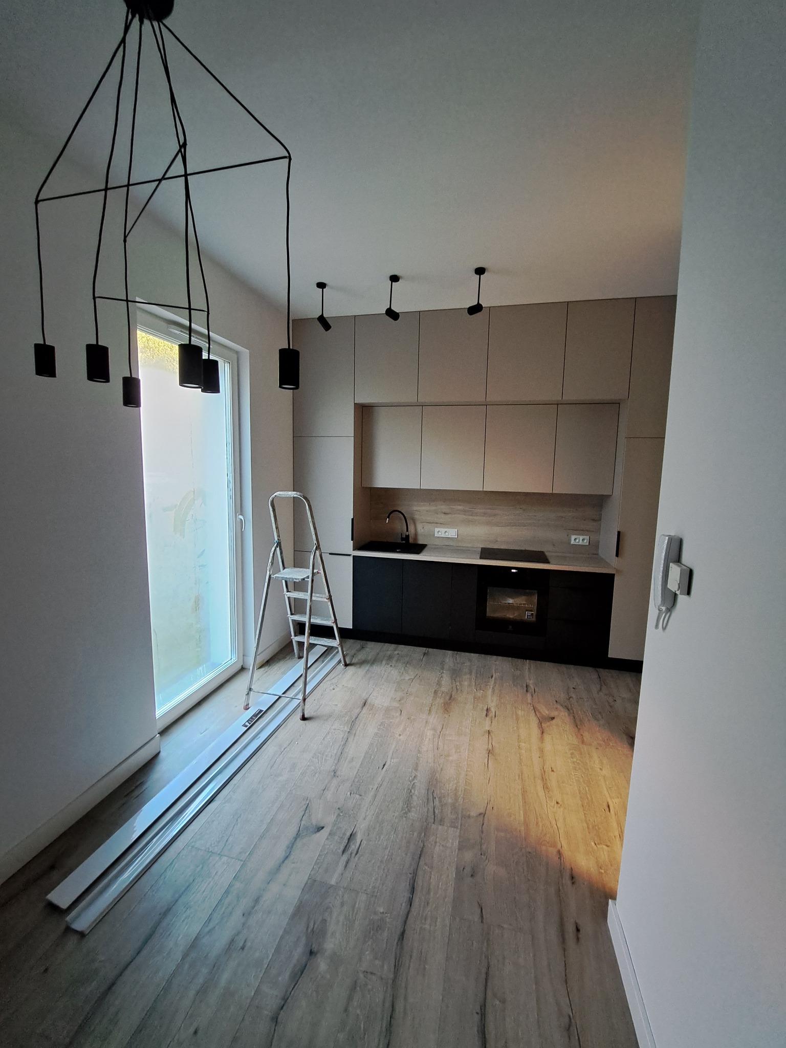 Wnętrze nowocześnie urządzonej kuchni z czarnymi szafkami dolnymi i beżowymi górnymi, widoczne elementy oświetlenia: wisząca lampa nad oknem i reflektory sufitowe, na podłodze panele imitujące...