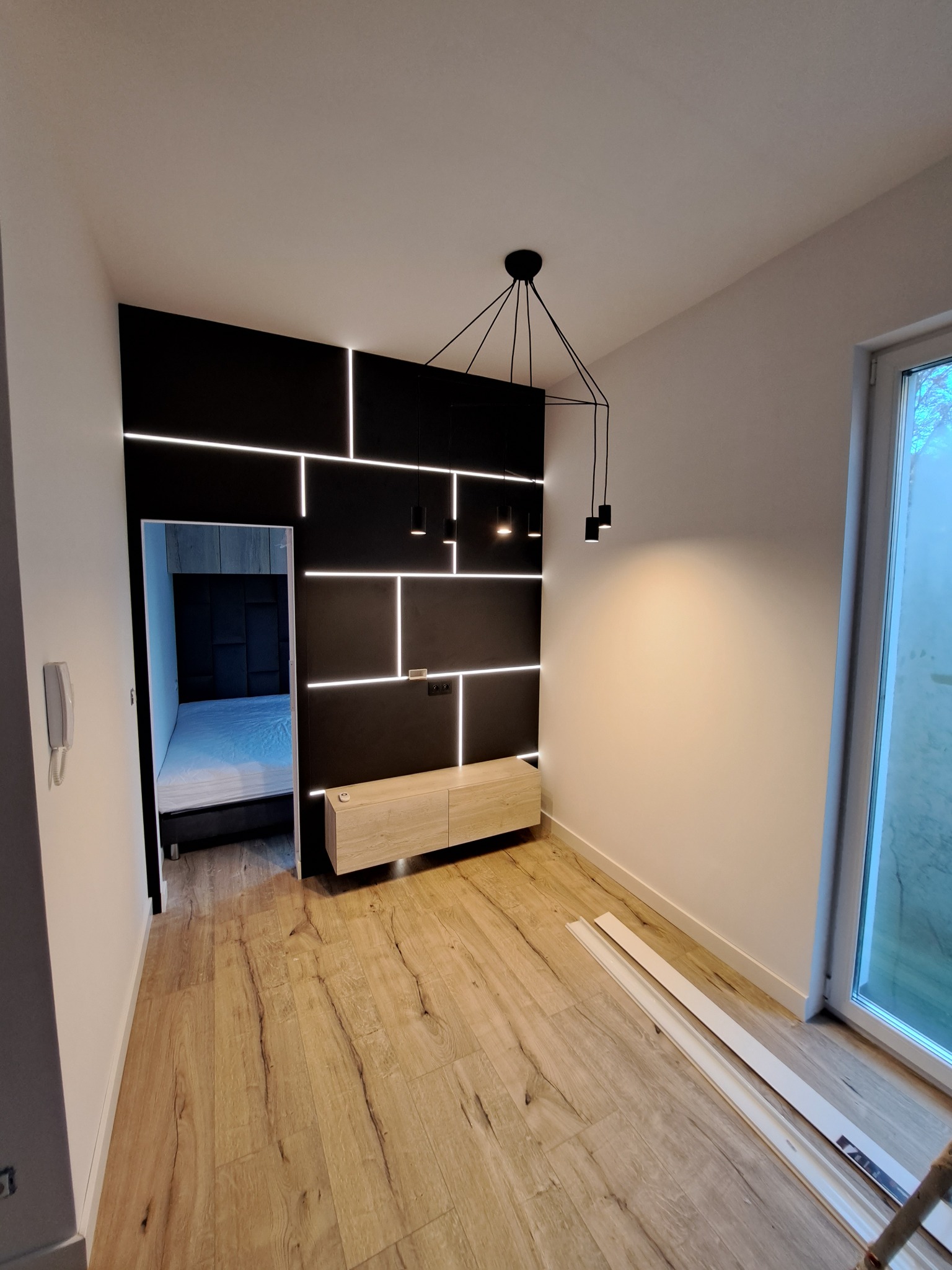 Nowoczesny salon z czarną ścianą dekoracyjną podświetloną ledami, drewnianą podłogą i wiszącą lampą w stylu industrialnym. W tle widoczne wejście do sypialni.