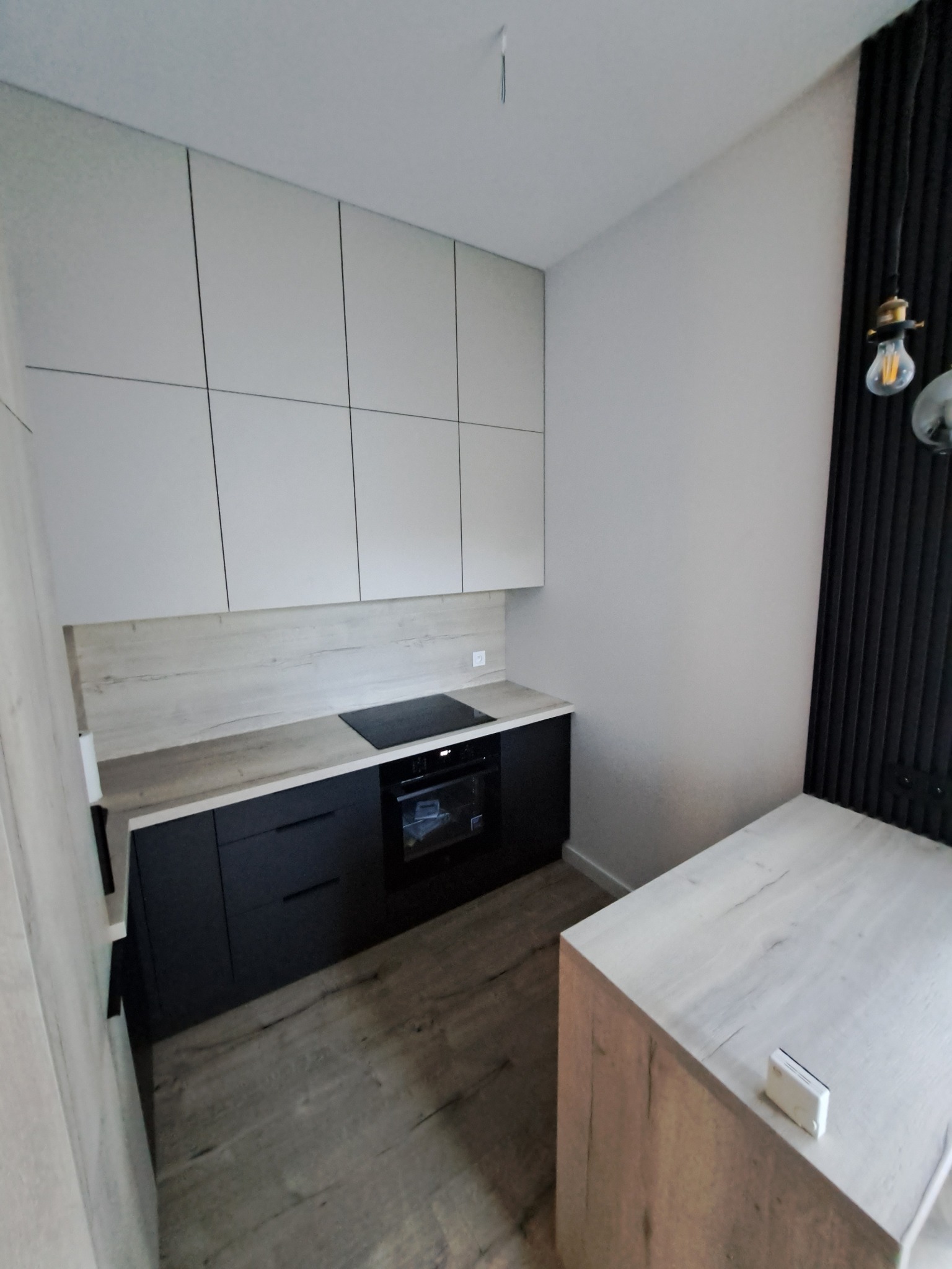 Nowoczesna kuchnia z białymi szafkami górnymi, czarnymi szafkami dolnymi i blatem imitującym drewno, z widoczną płytą indukcyjną i piekarnikiem. Na podłodze panele podłogowe imitujące drewno.