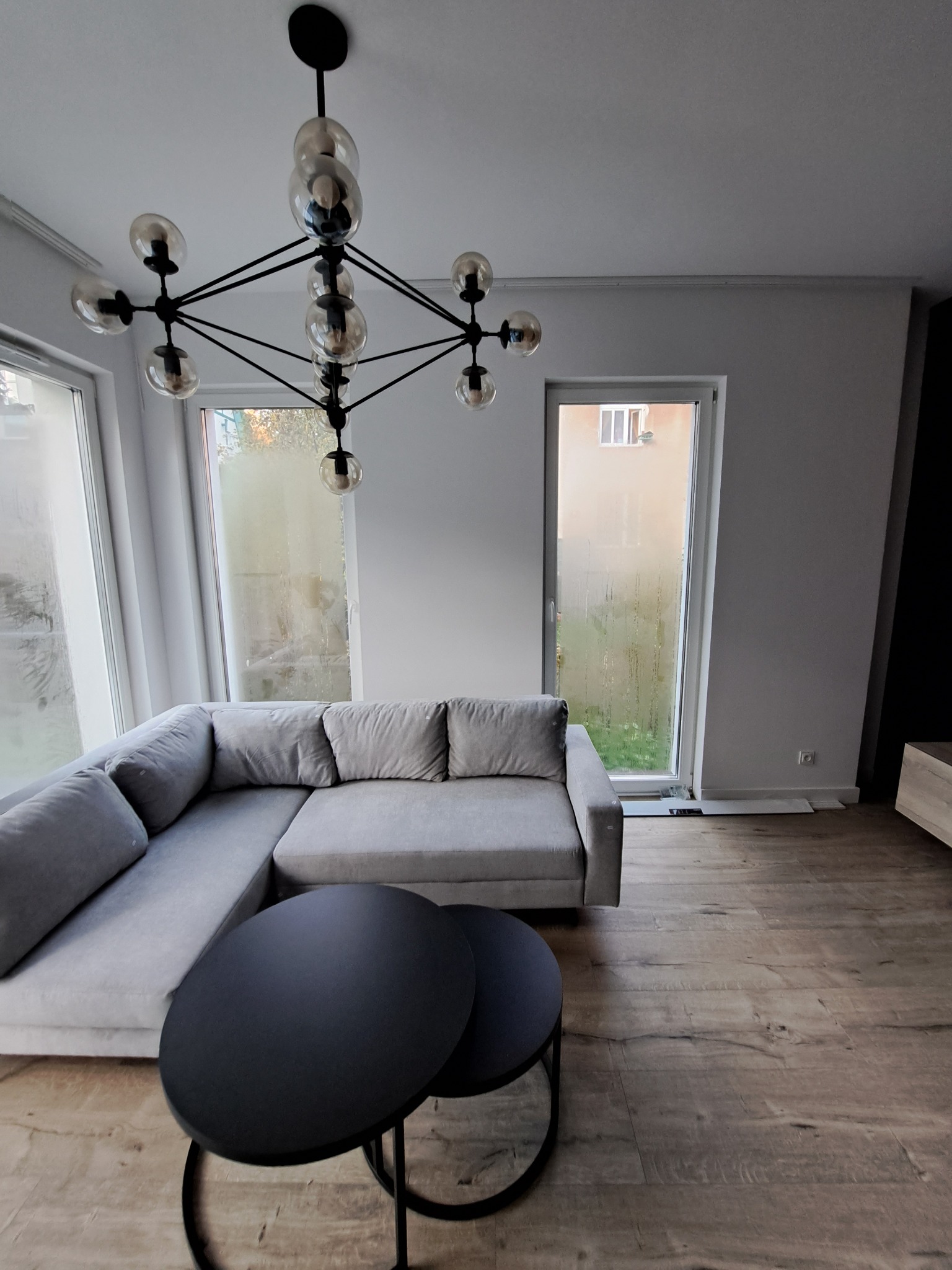 Jasne wnętrze salonu z szarą sofą narożną, okrągłymi stolikami kawowymi i nowoczesną lampą sufitową z kulistymi kloszami, parkiet na podłodze.