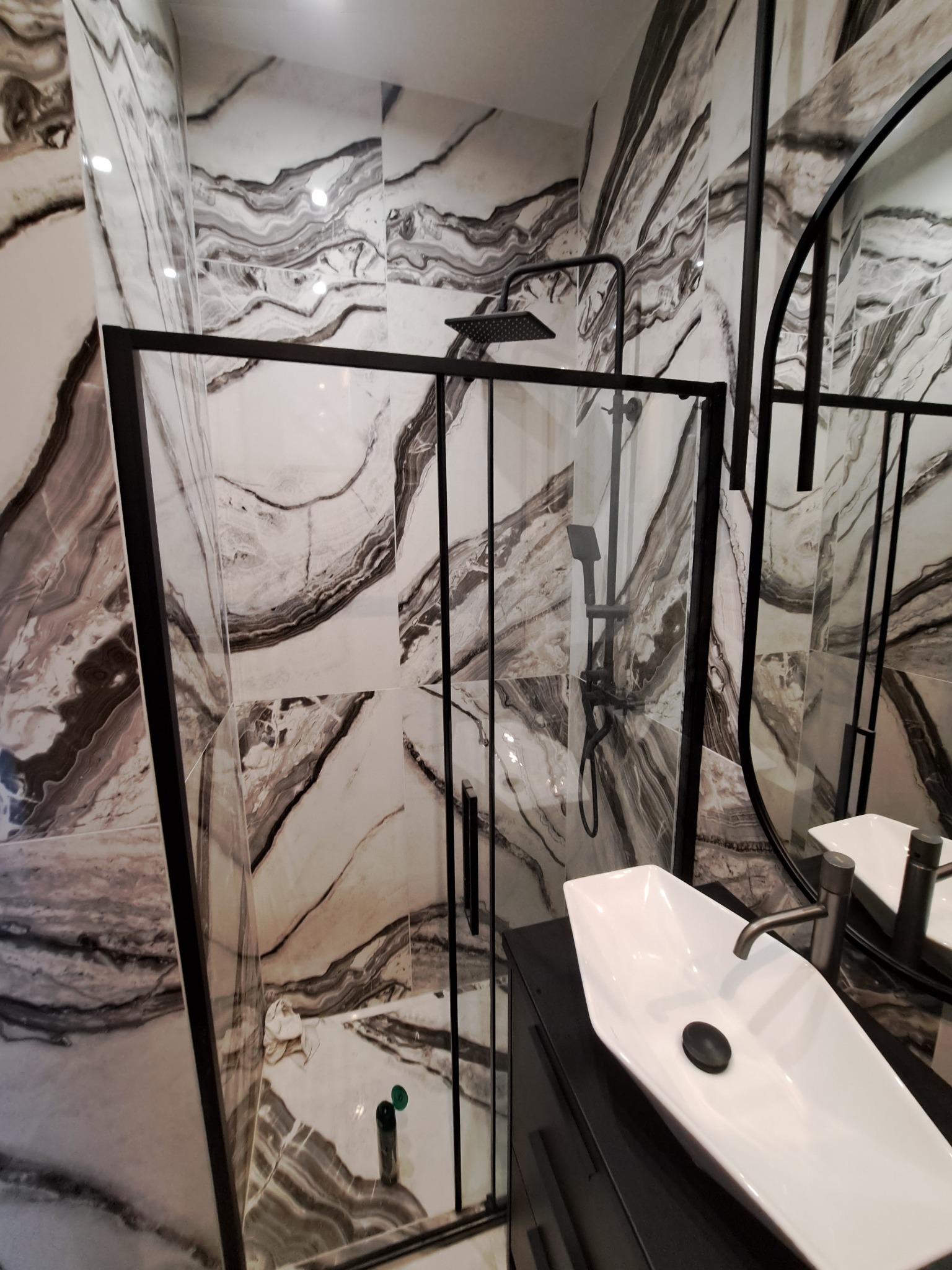 Nowoczesna łazienka z kabiną prysznicową typu walk-in z czarnymi profilami, umywalką nablatową o nietypowym kształcie i marmurowymi płytkami na ścianach.