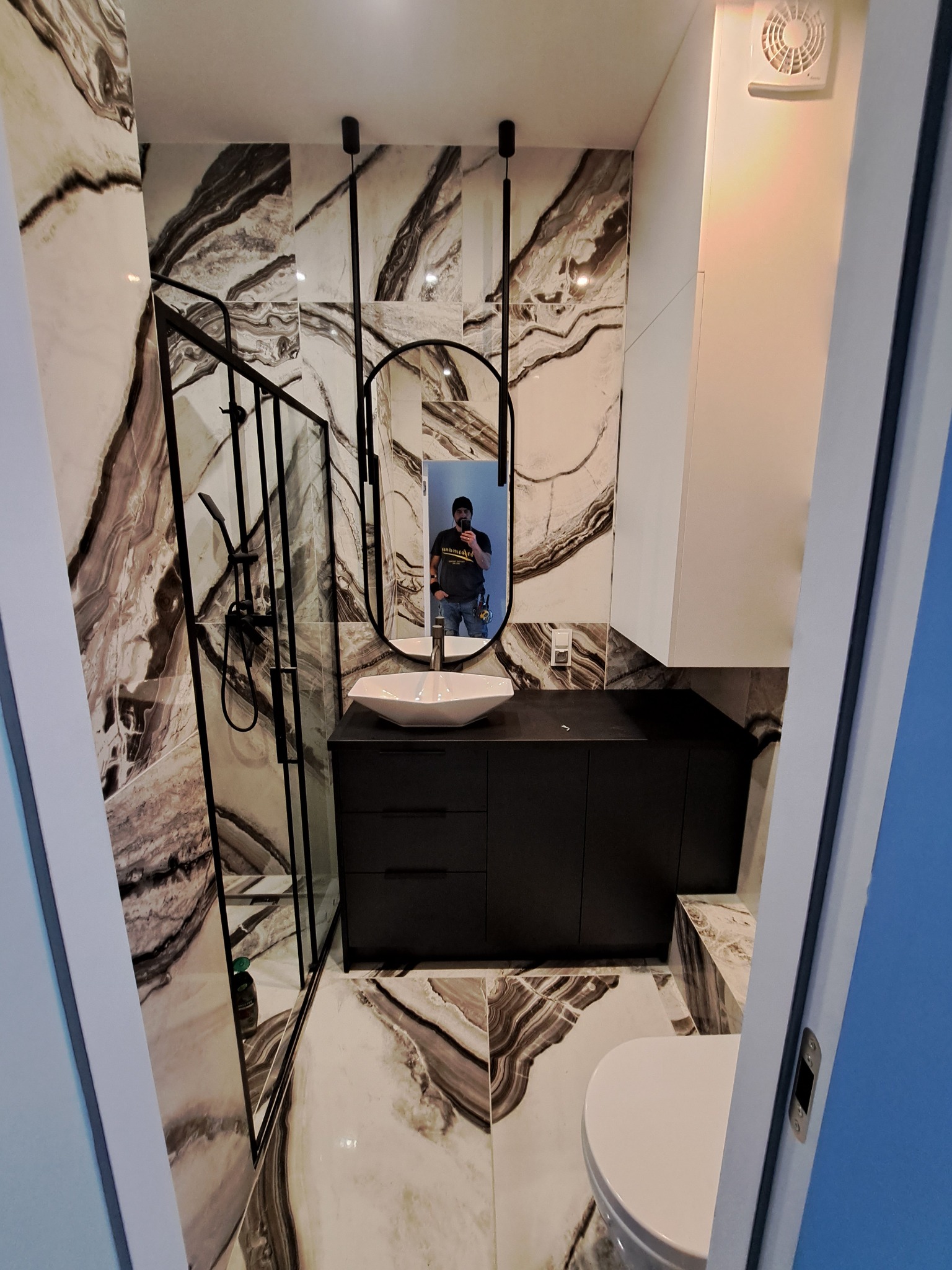 Nowoczesna łazienka z marmurowymi płytkami, czarną armaturą i kabiną prysznicową typu walk-in, widok zza otwartych drzwi.