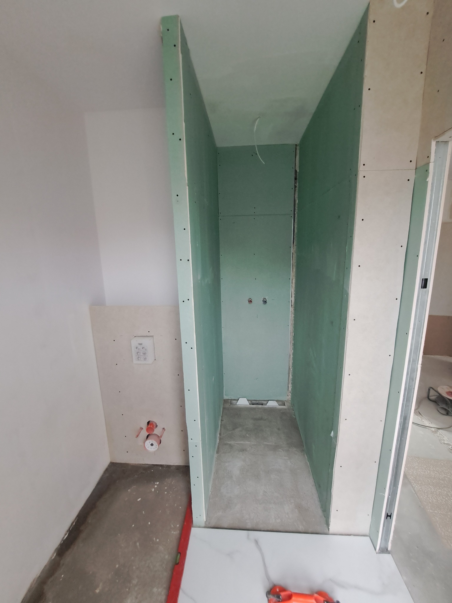 Łazienka w trakcie budowy: widoczne płyty kartonowo-gipsowe tworzące ścianki działowe, przygotowane instalacje wodne i elektryczne, oraz częściowo ułożone białe płytki podłogowe, poziomica...