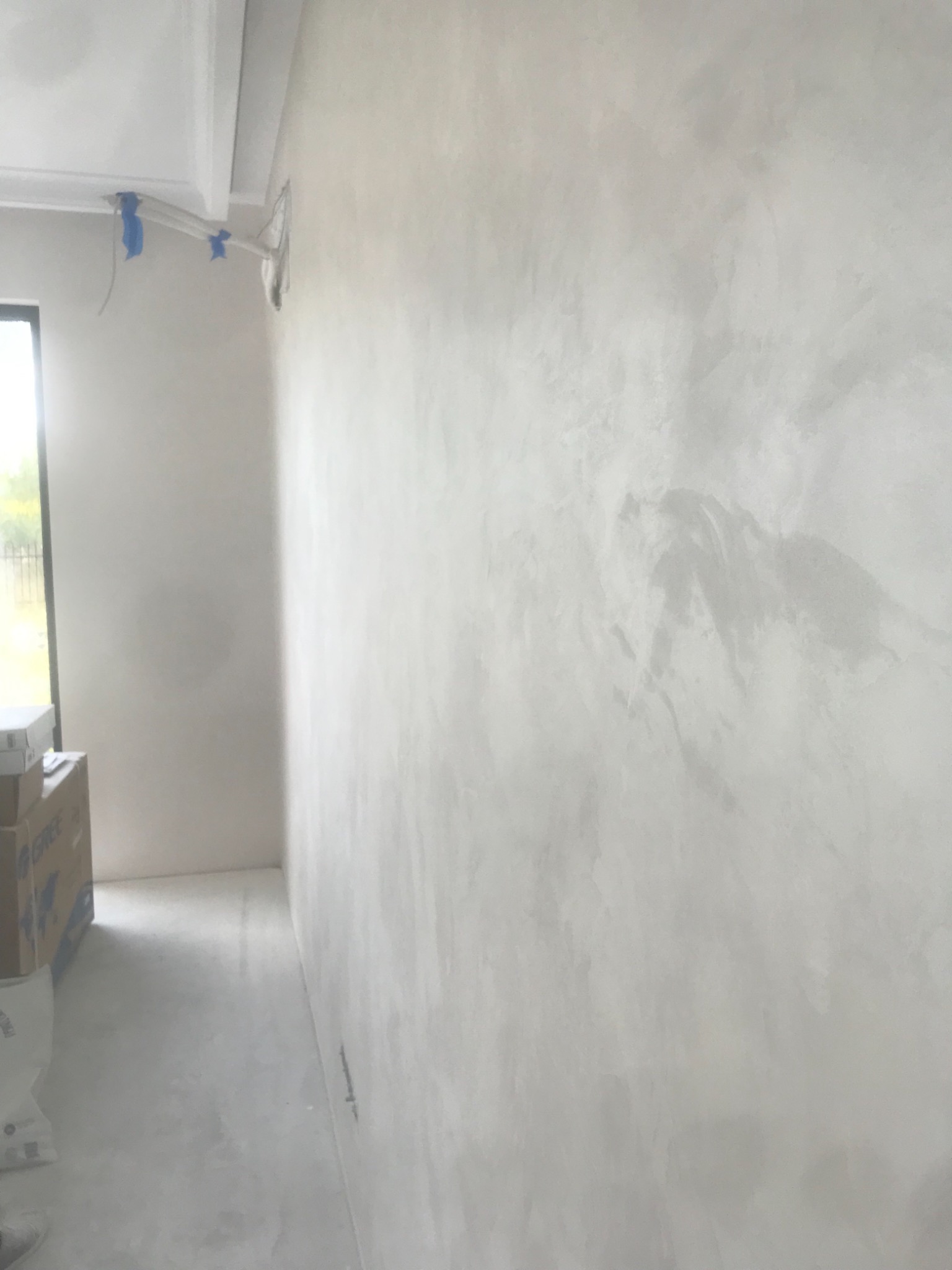 Ściana pokryta dekoracyjną, jasnoszarą gładzią w stylu weneckim, z widocznymi smugami i refleksami światła, w tle okno i kartony.