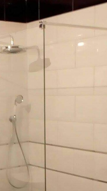 Szklana kabina prysznicowa z deszczownicą i słuchawką, wykończona białymi, prostokątnymi płytkami z szarą fugą, widoczny chromowany zawias mocujący szybę.