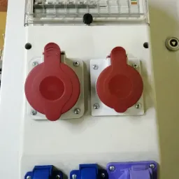 Rozdzielnica elektryczna z zabezpieczeniami nad gniazdami przemysłowymi z klapkami w kolorach czerwonym, niebieskim i fioletowym, zamontowana na białej obudowie.