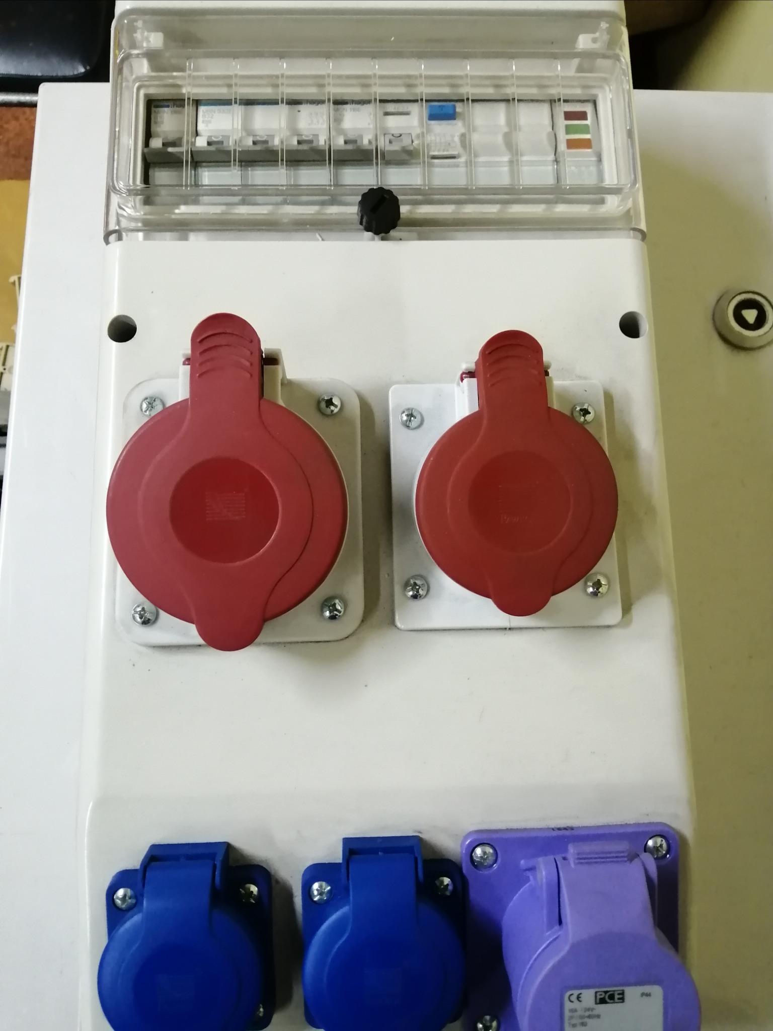 Rozdzielnica elektryczna z zabezpieczeniami nad gniazdami przemysłowymi z klapkami w kolorach czerwonym, niebieskim i fioletowym, zamontowana na białej obudowie.