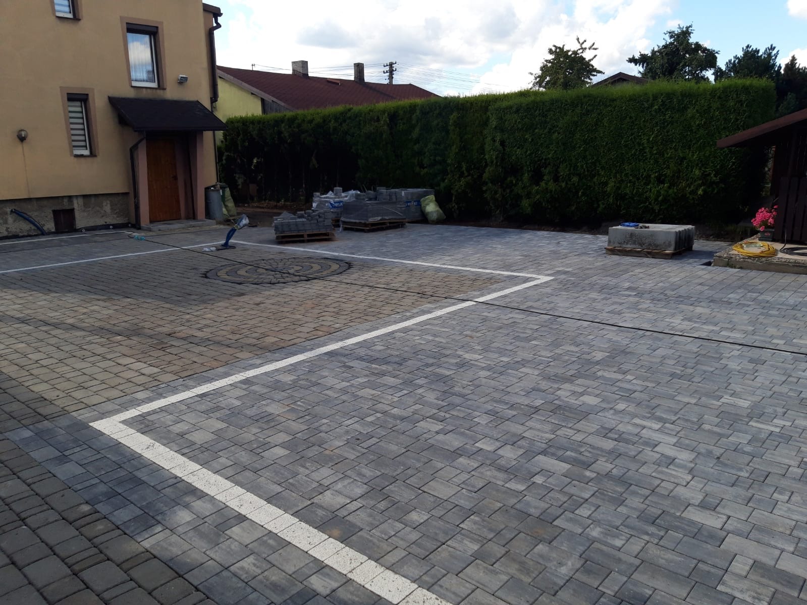 Ukończona nawierzchnia z szarej kostki brukowej z wyznaczonymi białymi liniami parkingowymi, w tle dom z brązowymi drzwiami i zielony żywopłot.