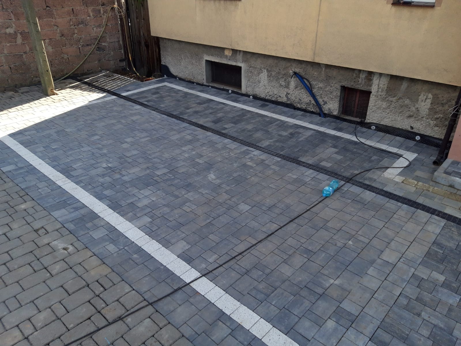 Ułożona kostka brukowa w odcieniach szarości z białymi pasami oddzielającymi fragmenty. Widoczne czarne korytka odwadniające wzdłuż ściany budynku i na granicy kostki. Na kostce leży niebieska...