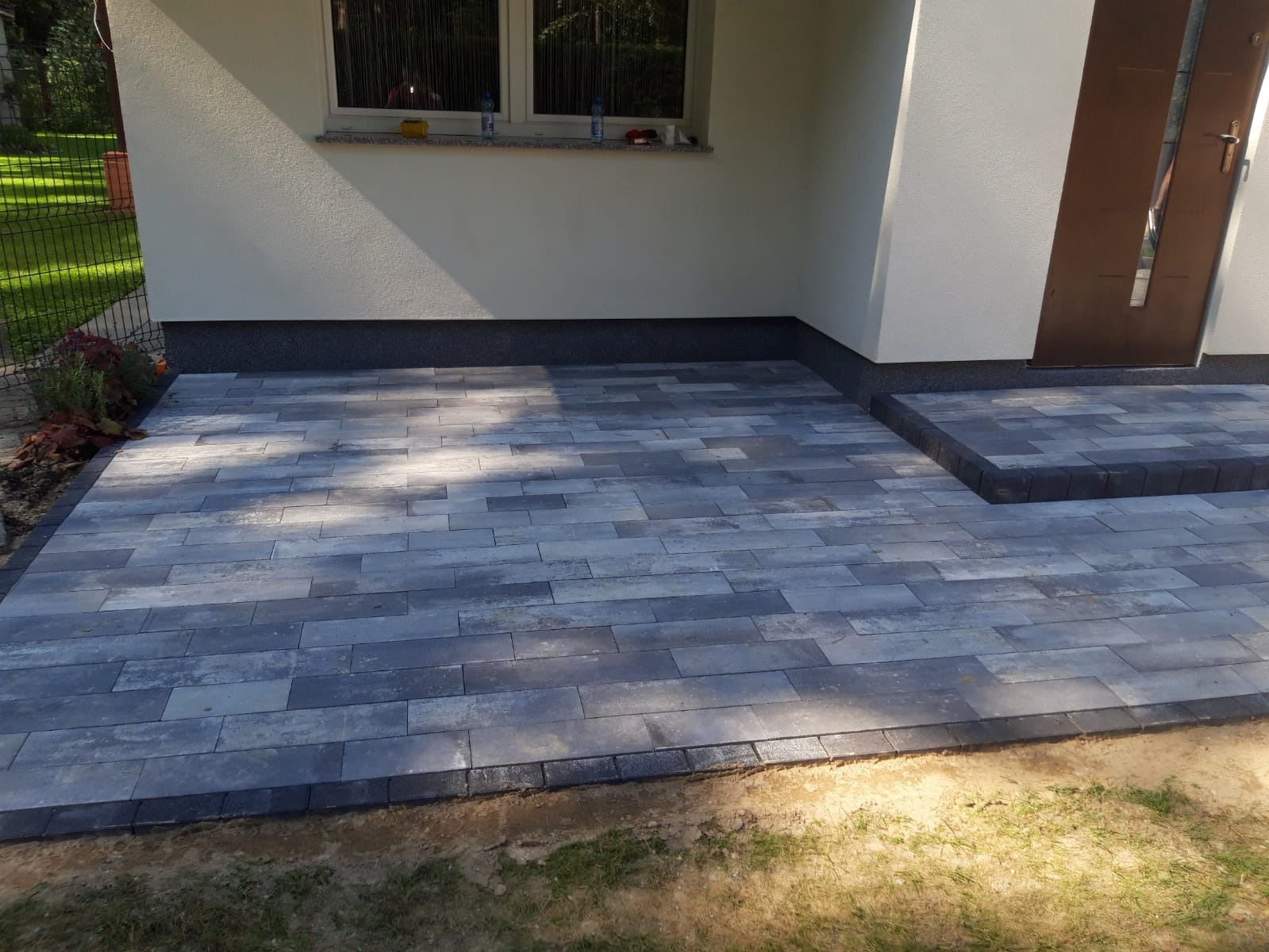 Ułożona szara kostka brukowa w kształcie prostokątów tworząca chodnik przed budynkiem z widocznymi schodami wejściowymi, obrzeża chodnika wykończone ciemniejszą kostką.