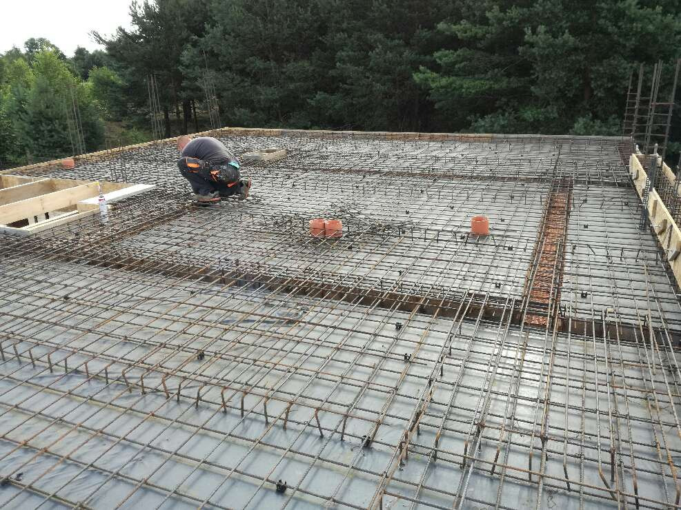 Zbrojenie płyty fundamentowej z pracownikiem w trakcie montażu, widoczne rury kanalizacyjne i elementy drewniane szalunku w tle las.