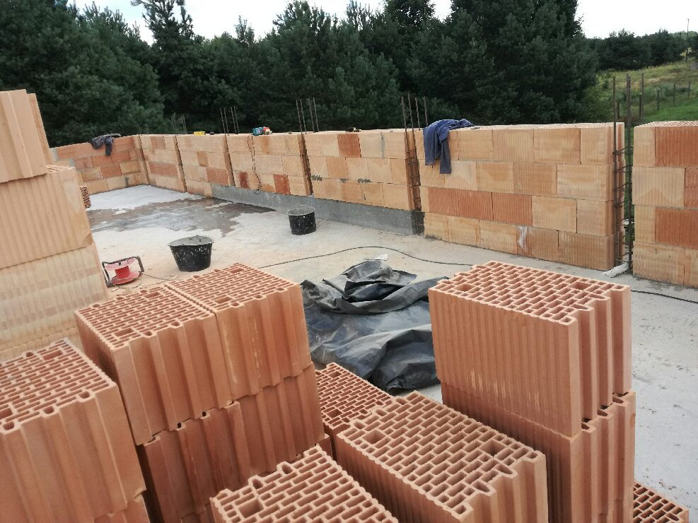 Wznoszenie ścian zewnętrznych budynku z bloczków ceramicznych Porotherm, widoczne zbrojenia pionowe, przygotowane zapasy materiału oraz narzędzia na placu budowy.