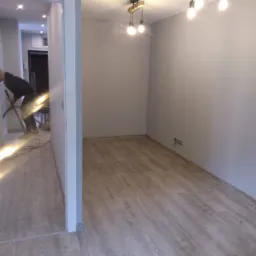 Pokój po remoncie z nową podłogą z paneli, widoczne lampy sufitowe z żarówkami i osoba odkurzająca w tle.