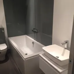 Nowoczesna łazienka z białą prostokątną wanną zabudowaną, umywalką nablatową na szafce z marmurowym blatem i szarą ścianą prysznicową ze szkła.