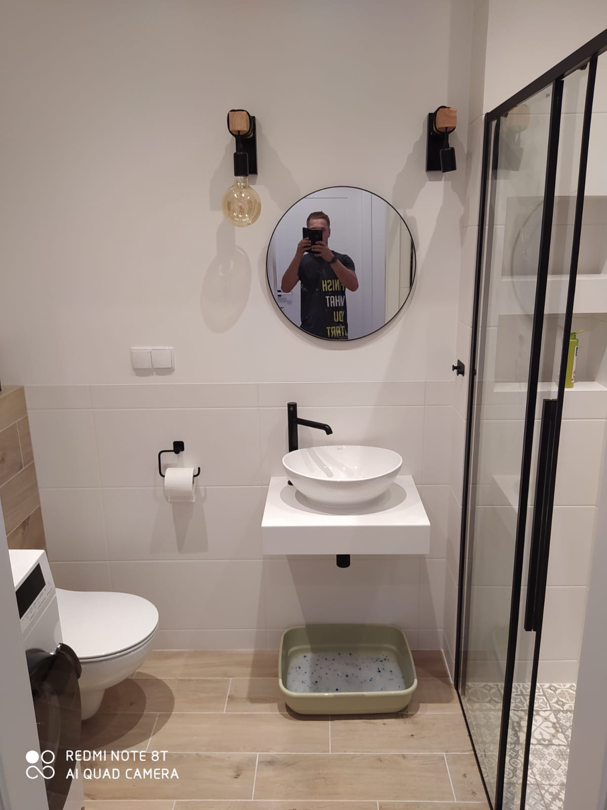 Nowoczesna łazienka z białą ceramiką, czarną armaturą i oświetleniem typu Edison, z odbiciem fotografa w okrągłym lustrze nad umywalką.