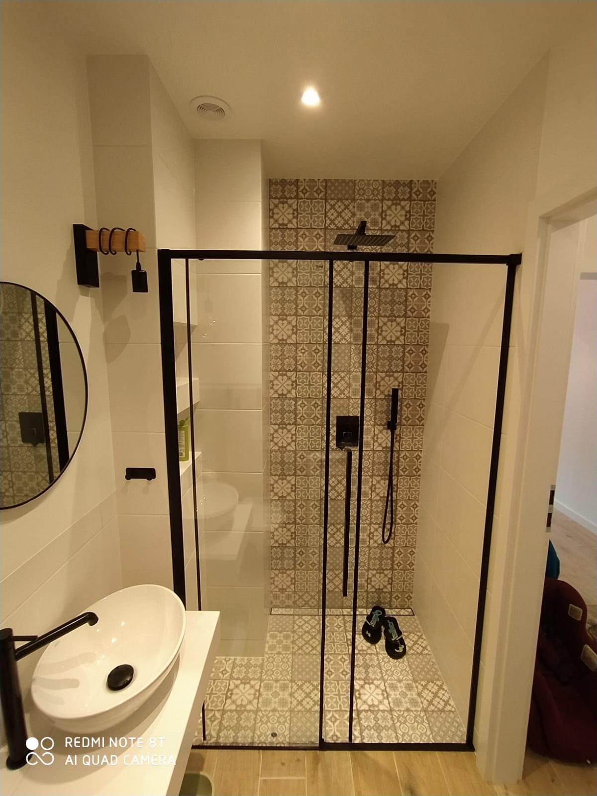 Nowoczesna łazienka po remoncie: kabina prysznicowa typu walk-in z czarnymi ramami, umywalka nablatowa owalna, lustro okrągłe, płytki patchworkowe pod prysznicem i na podłodze, drewniane wieszaki...